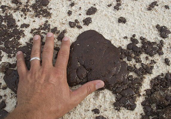 Hand near dark oil spill on sand.